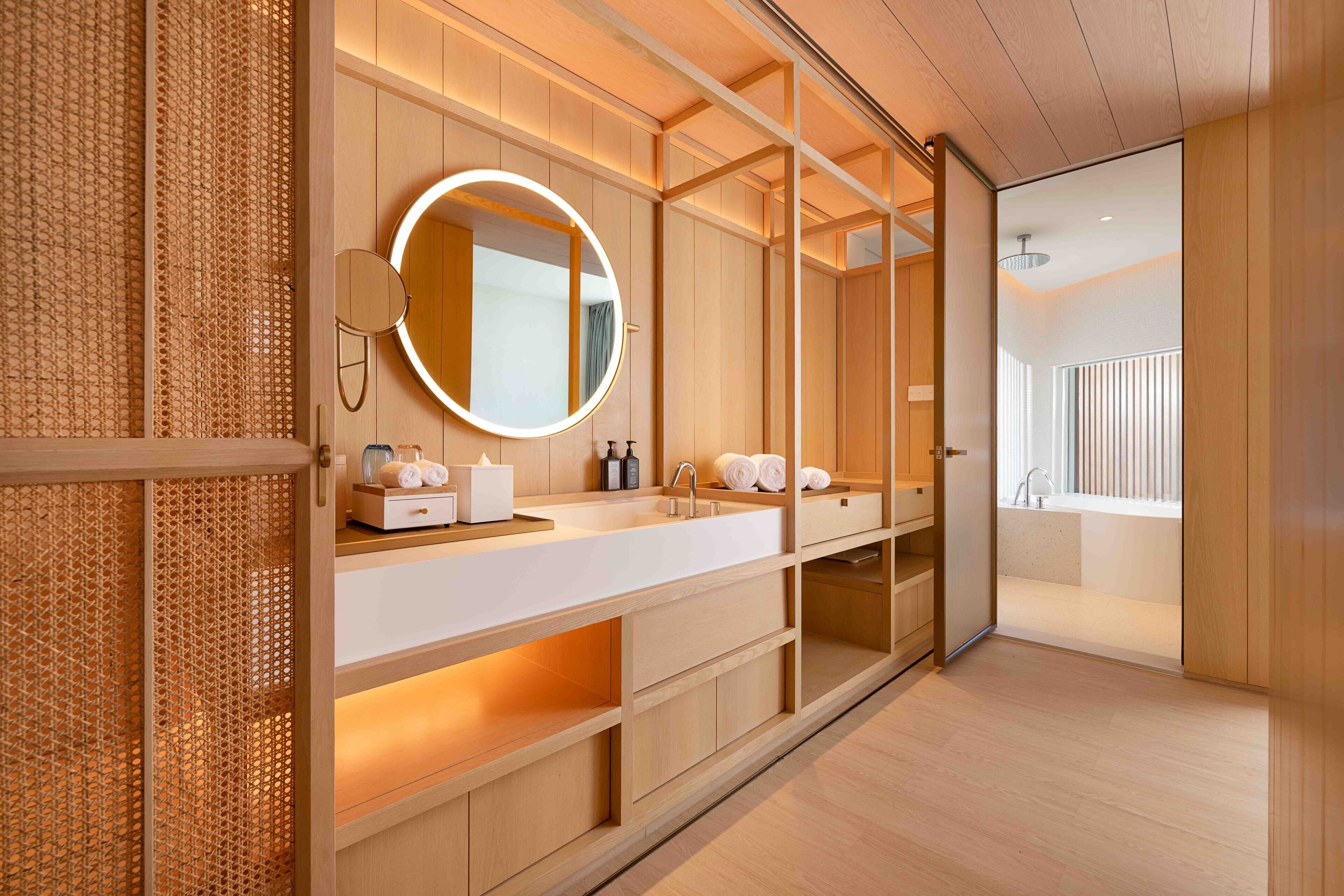 A modern bathroom with a wooden vanity, a round mirror with a light, and a view of a spa area.