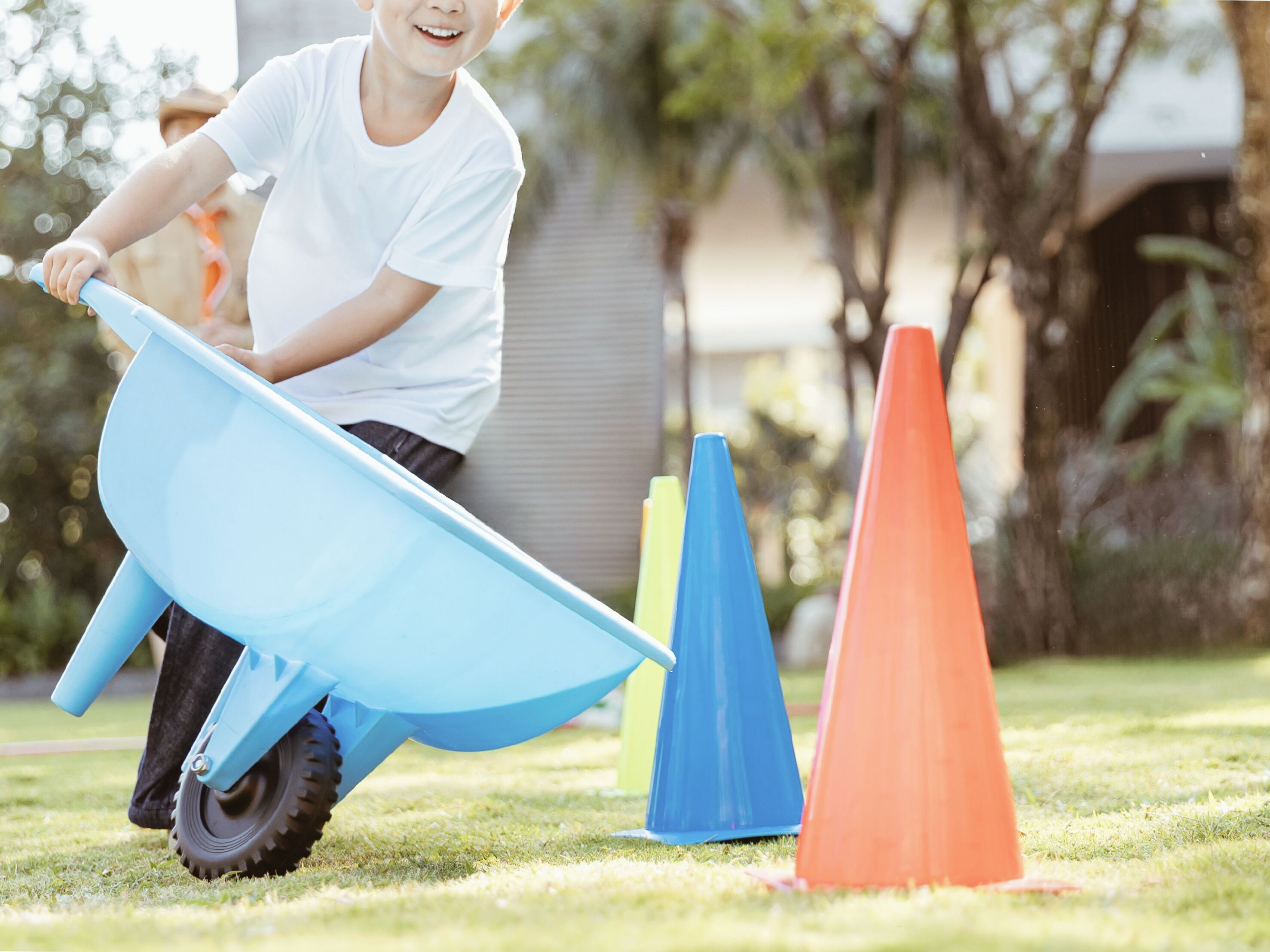 A child pushing a blue wheelbarrow on grass with colorful cones.