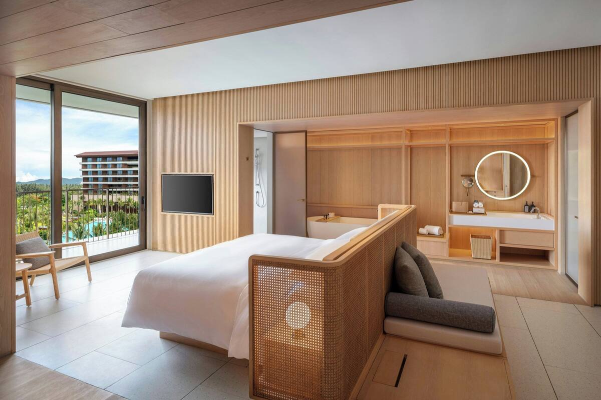 A modern hotel room with a large bed, a TV, a balcony with a view of a building and greenery, a bathroom with a mirror and sink, and a seating area.
