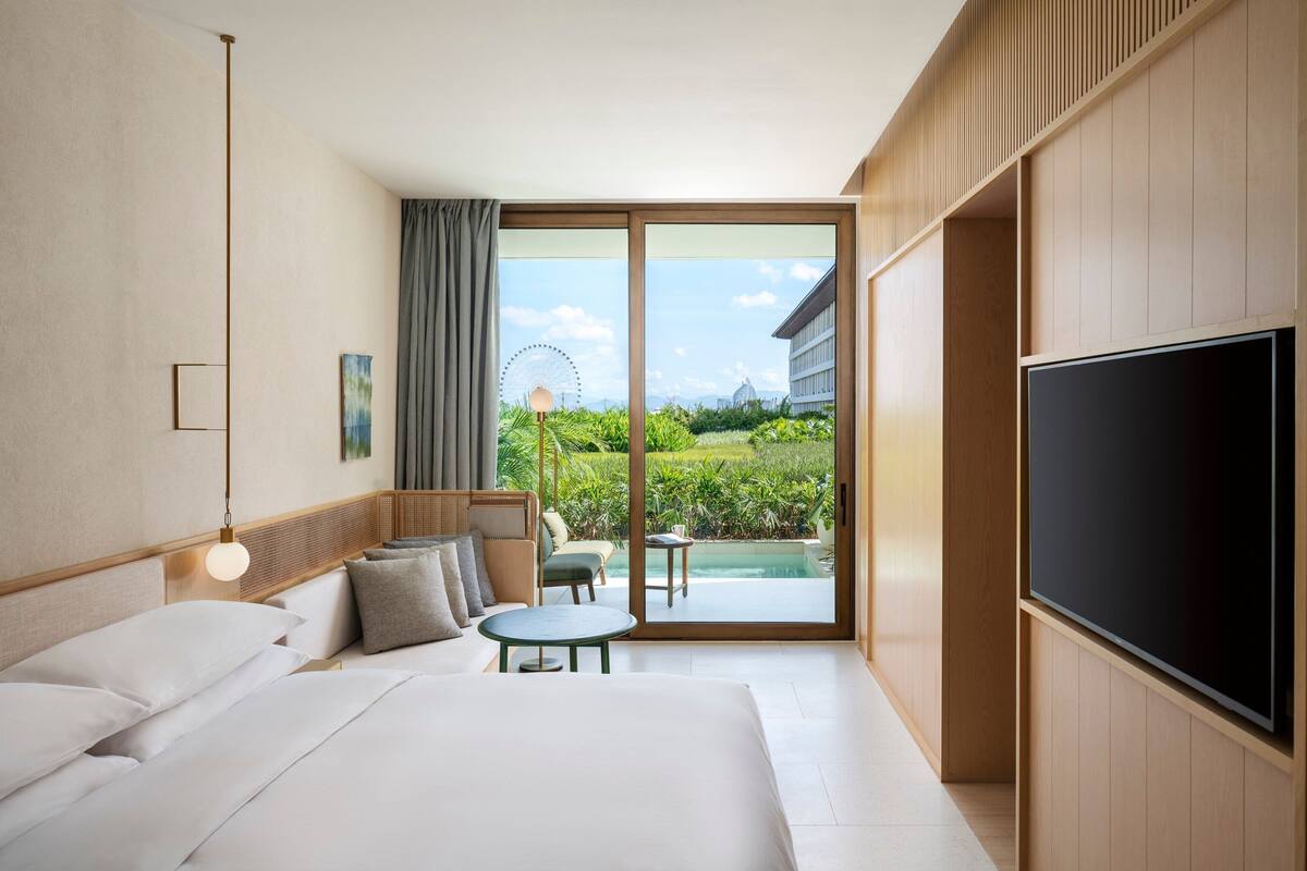 A hotel room with a bed, a TV, a seating area with a table, and a view of a pool and Ferris wheel.