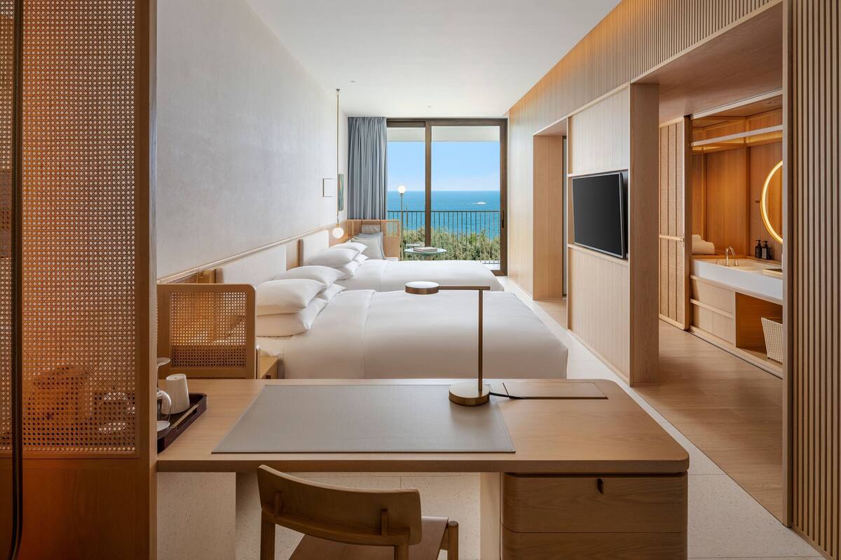 A modern hotel room with a large bed, a desk, a TV, and a balcony with a view of the sea.