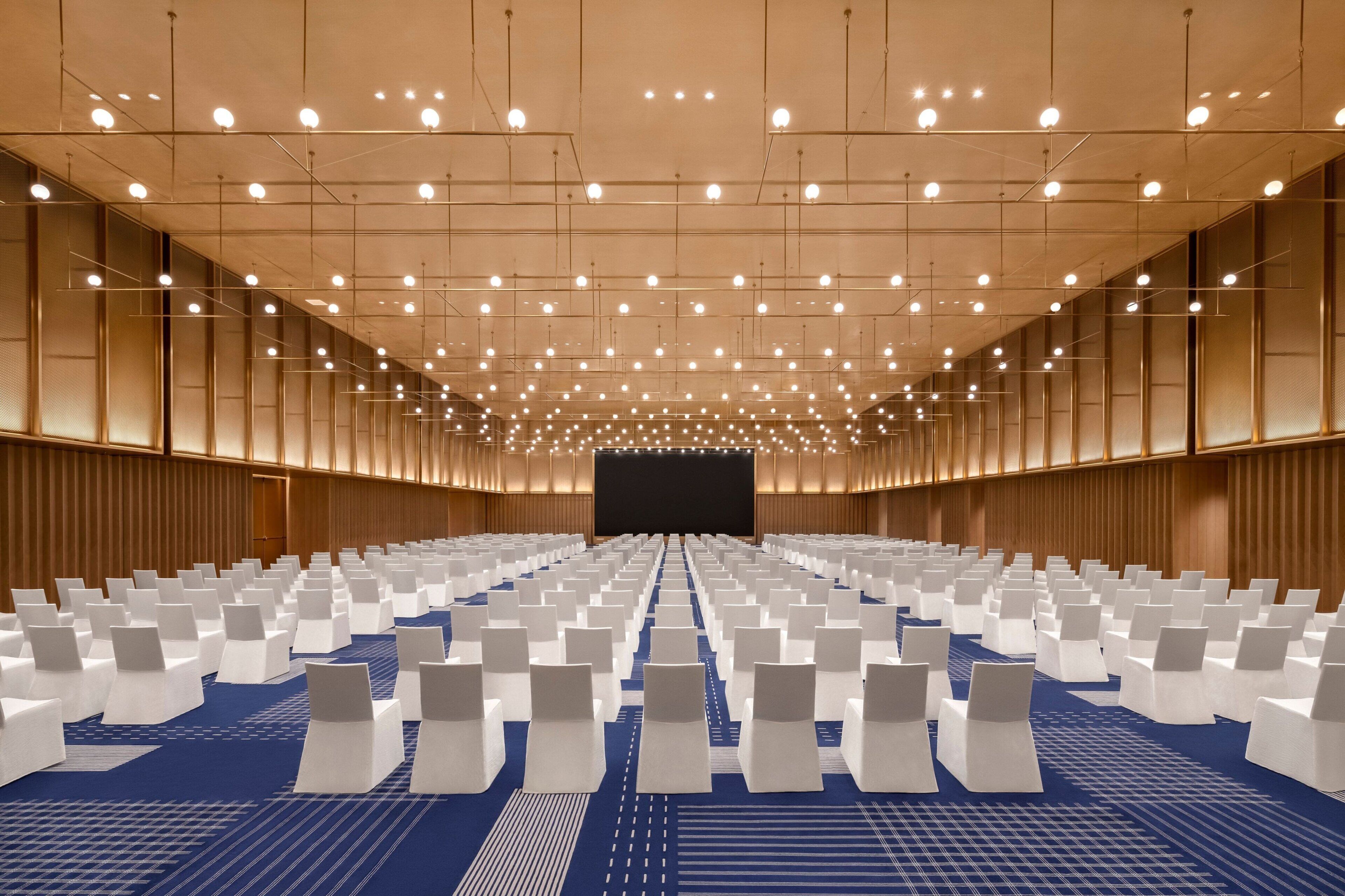 A large conference room with rows of white chairs, a blue carpet, and a stage at the front.