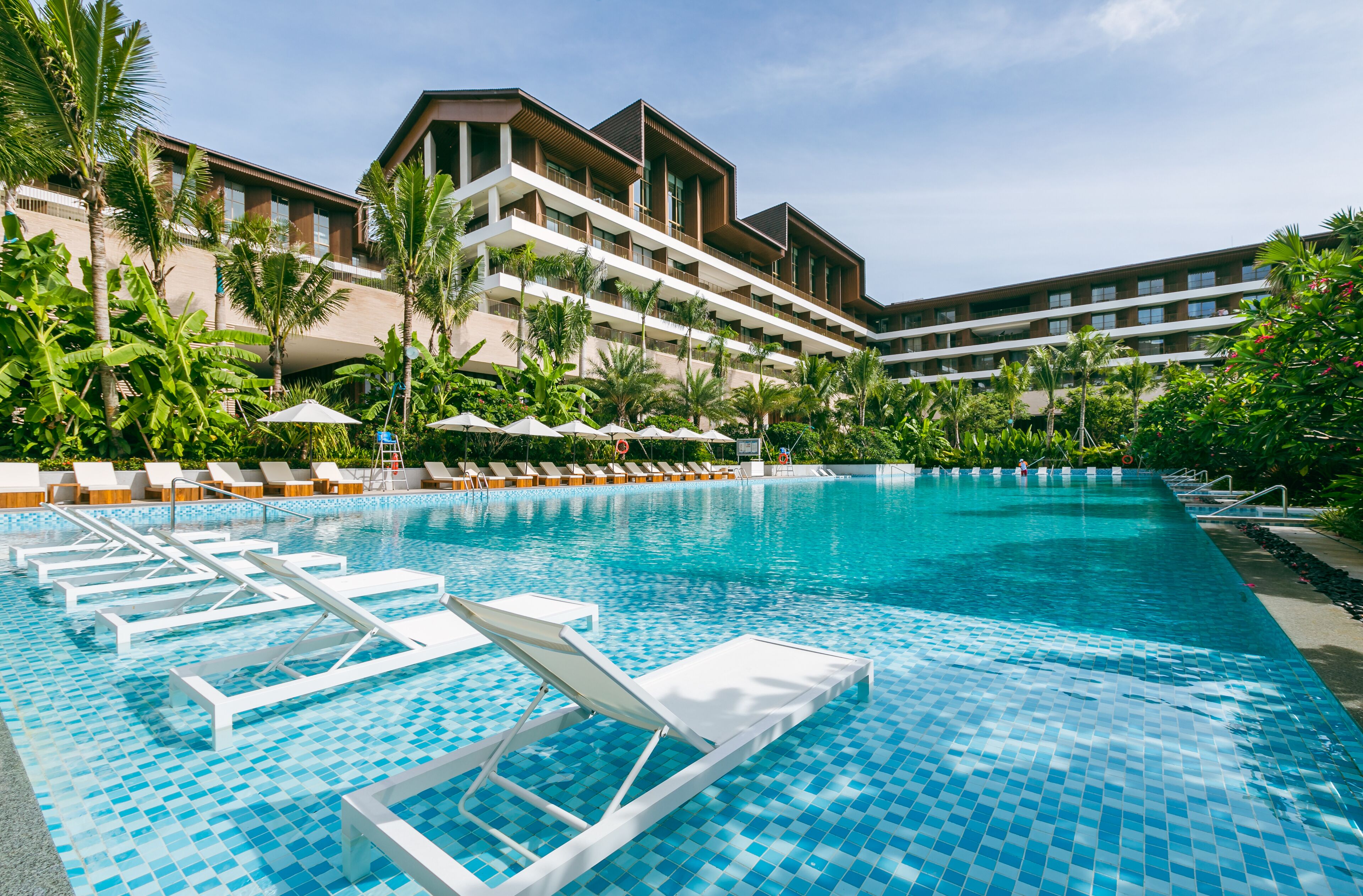 Una gran piscina con sillas de descanso y palmeras, rodeada por un edificio de hotel de varios pisos.