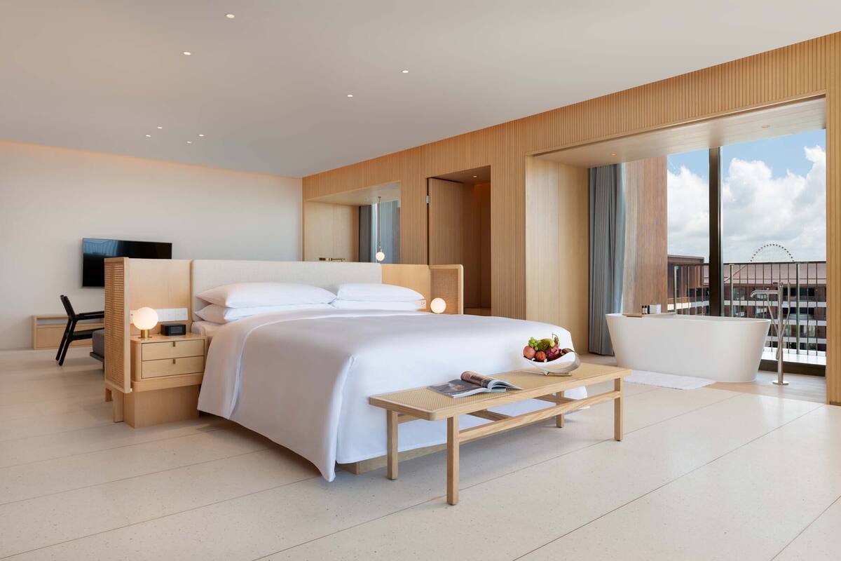 A modern hotel room with a large bed, bedside tables, a desk, a chair, a small table with a book and fruit, a bathtub, and large windows with a view of the city.