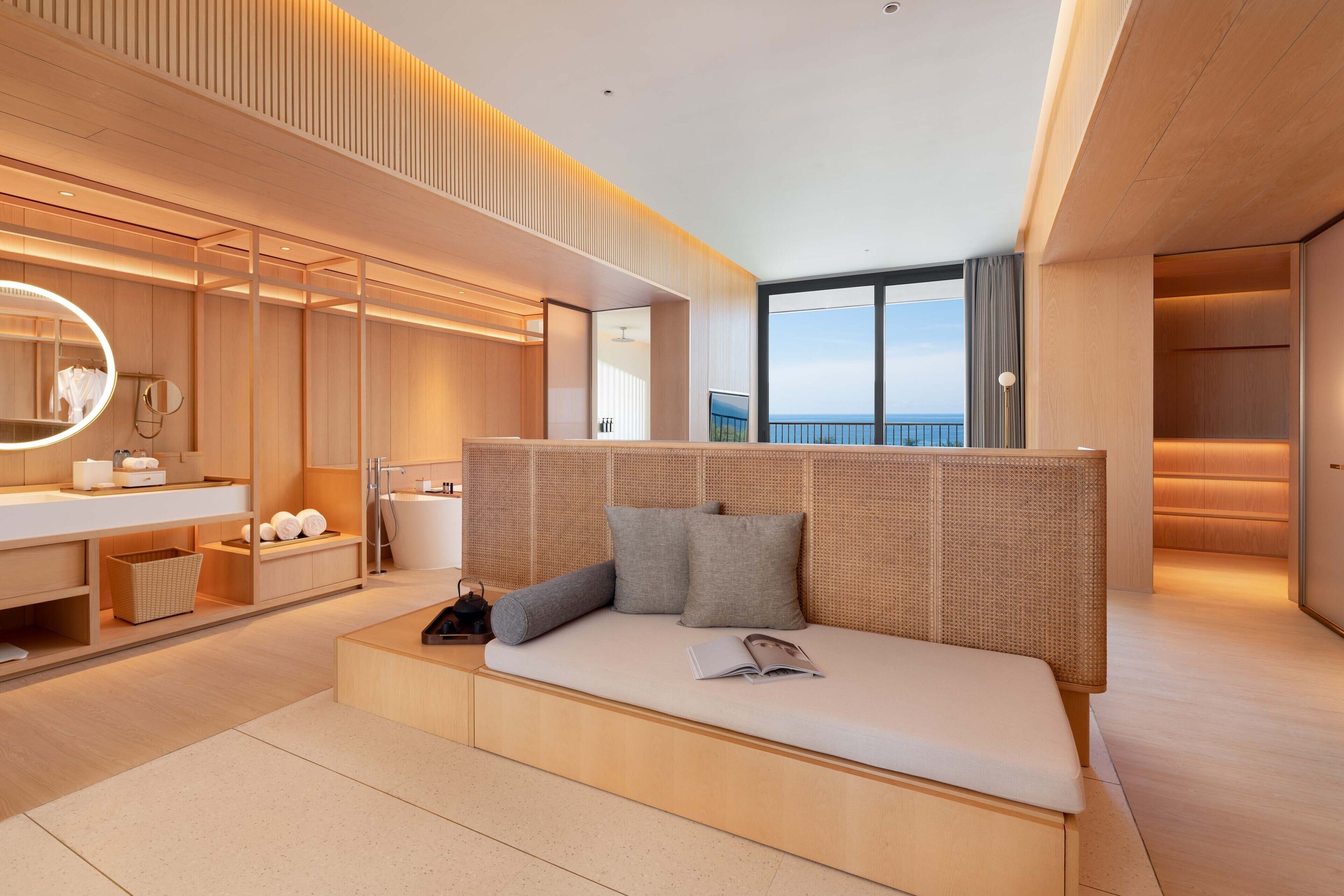A modern hotel room with a large bed, a built-in desk, a vanity with a mirror, and a view of the sea.