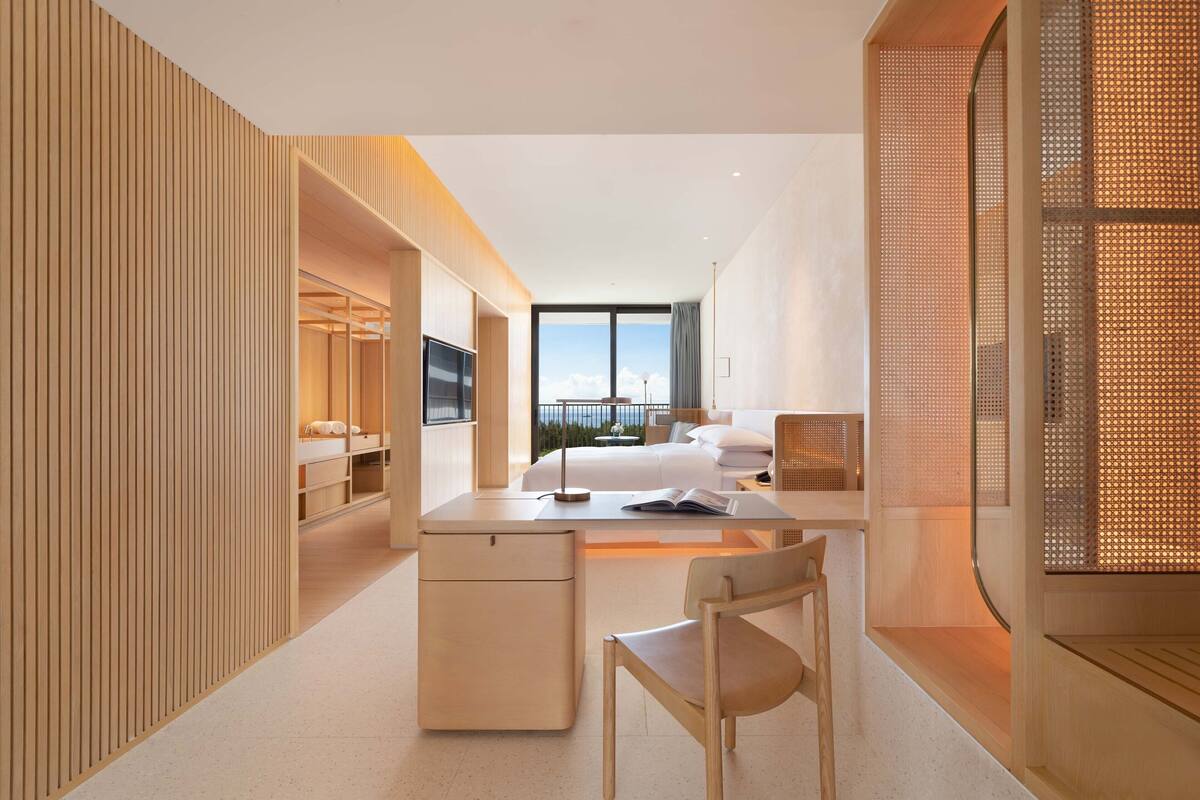 A modern hotel room with a large bed, a desk with a chair, a TV, and a view of the outdoors.
