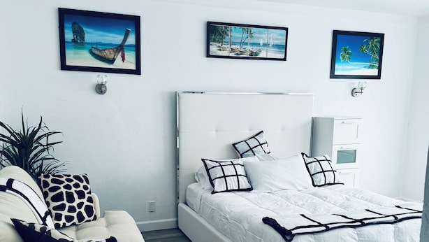 Een moderne slaapkamer met een wit bed, een beige bank en een kamerbreed tapijt met een patroon. Drie ingekaderde foto's hangen aan de muur.