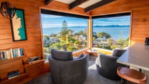 A room with large windows offering a view of a coastal landscape, a wooden bookshelf, and two grey armchairs. - Roydons - Kaiteriteri Upstairs Holiday Unit (Kaiteriteri)
