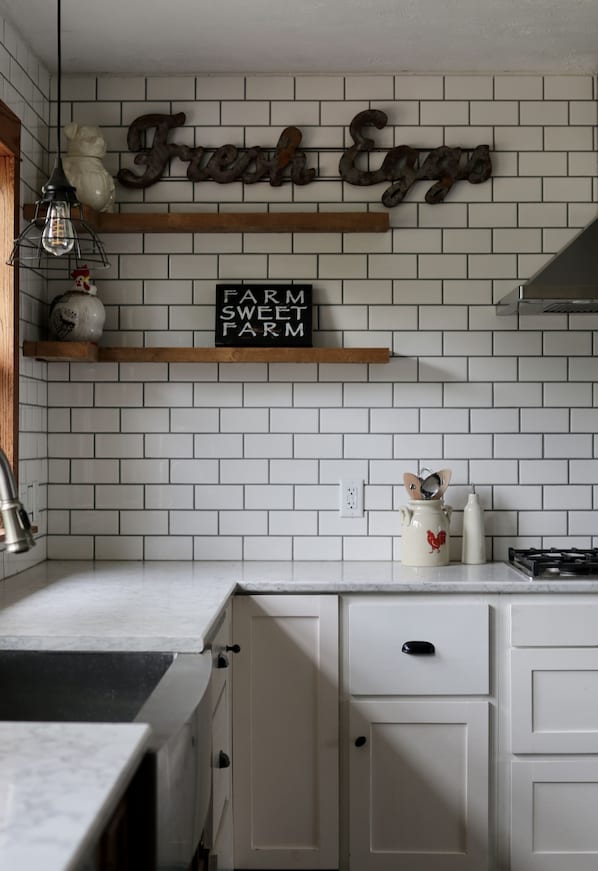 A kitchen with white tiled walls, a wooden shelf with a sign reading "Farm Sweet Farm", and a decorative cat mug on the counter.