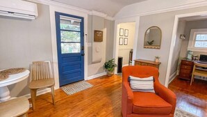 A room with a blue door, a wooden floor, a small table with chairs, a red armchair, and a desk with a computer.
