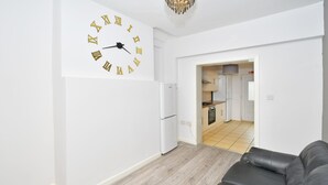 A modern living room with a black leather sofa, a white wall clock, and a kitchen visible through an open doorway. - Townhouse @ 115 Walthall Street Crewe (Crewe)