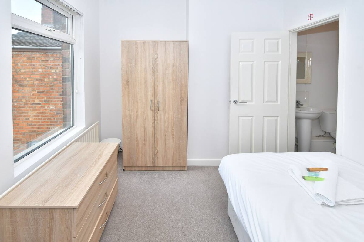 Une chambre à coucher avec une armoire en bois, un lit blanc et une fenêtre donnant sur un immeuble en briques.