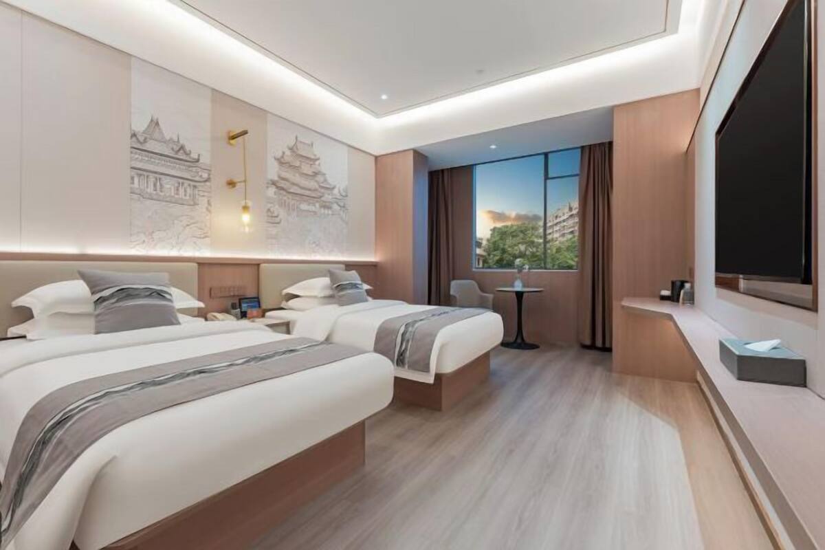 A hotel room with two beds, a small table, a chair, a TV, and artwork on the walls.