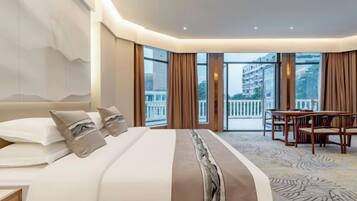 A hotel room with a large bed, a dining table, and a view of the city through large windows.