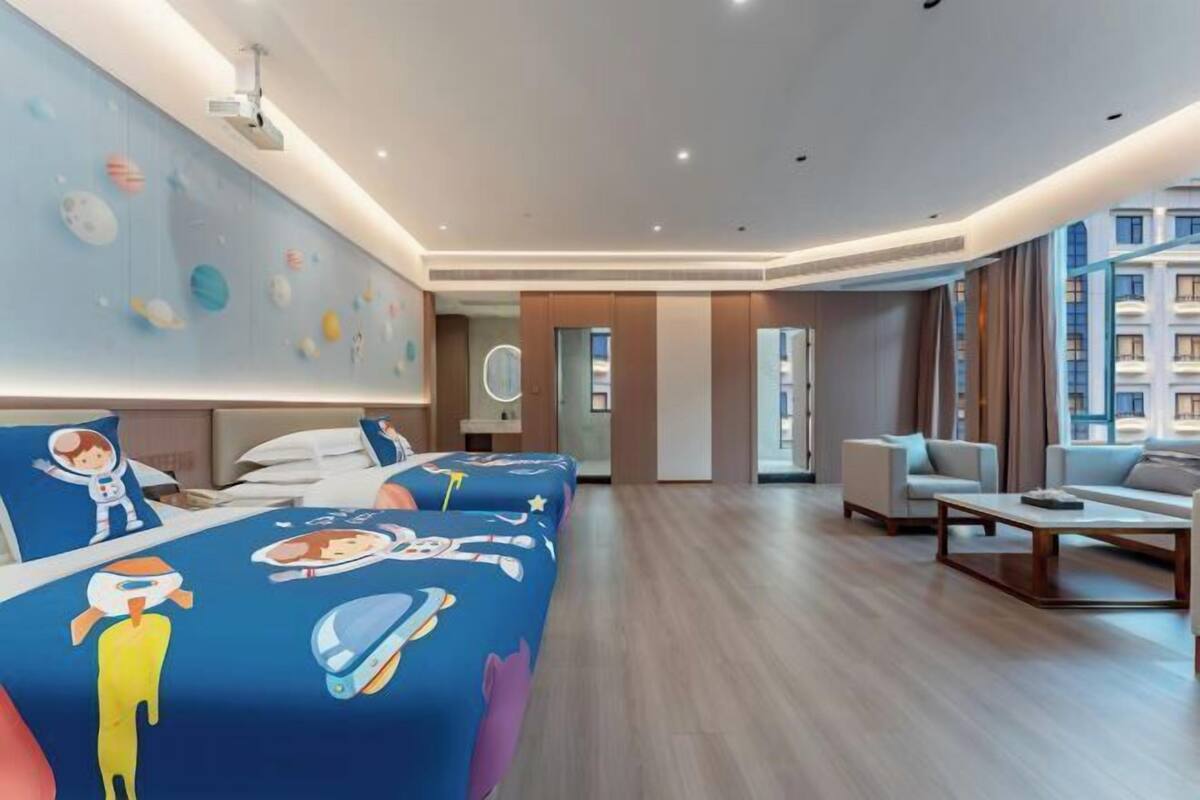A hotel room with a bed, a desk, a chair, a sofa, a coffee table, a wall with a space-themed mural, and a view of a building.