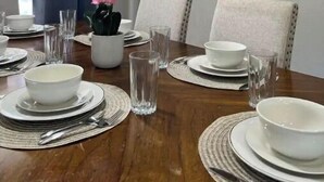 Un coin repas avec une table en bois dressée pour quatre personnes, des assiettes blanches, des bols et des verres transparents. Une orchidée en pot est posée sur la table, et un mur est décoré de dessous de verre en osier.