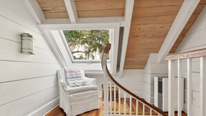 Une chambre mansardée chaleureuse avec un plafond en bois, une chaise blanche recouverte d’une couverture, une fenêtre donnant sur les arbres et un appareil de climatisation blanc.