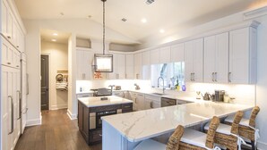 A modern kitchen with white cabinetry, a central island with bar stools, and a built-in dishwasher. - Starfish Cottage & Carriage House | Large Property with Private Pool (Rosemary Beach)