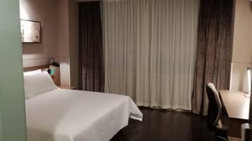 A hotel room with a bed, a desk, a chair, a nightstand, and a window with curtains.