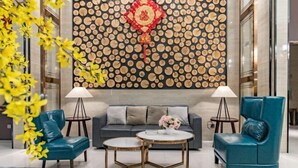 Um lobby moderno com um sofá cinzento, poltronas azul-turquesa e mesas redondas. É visível uma parede decorativa com padrões circulares e um ornamento pendente.
