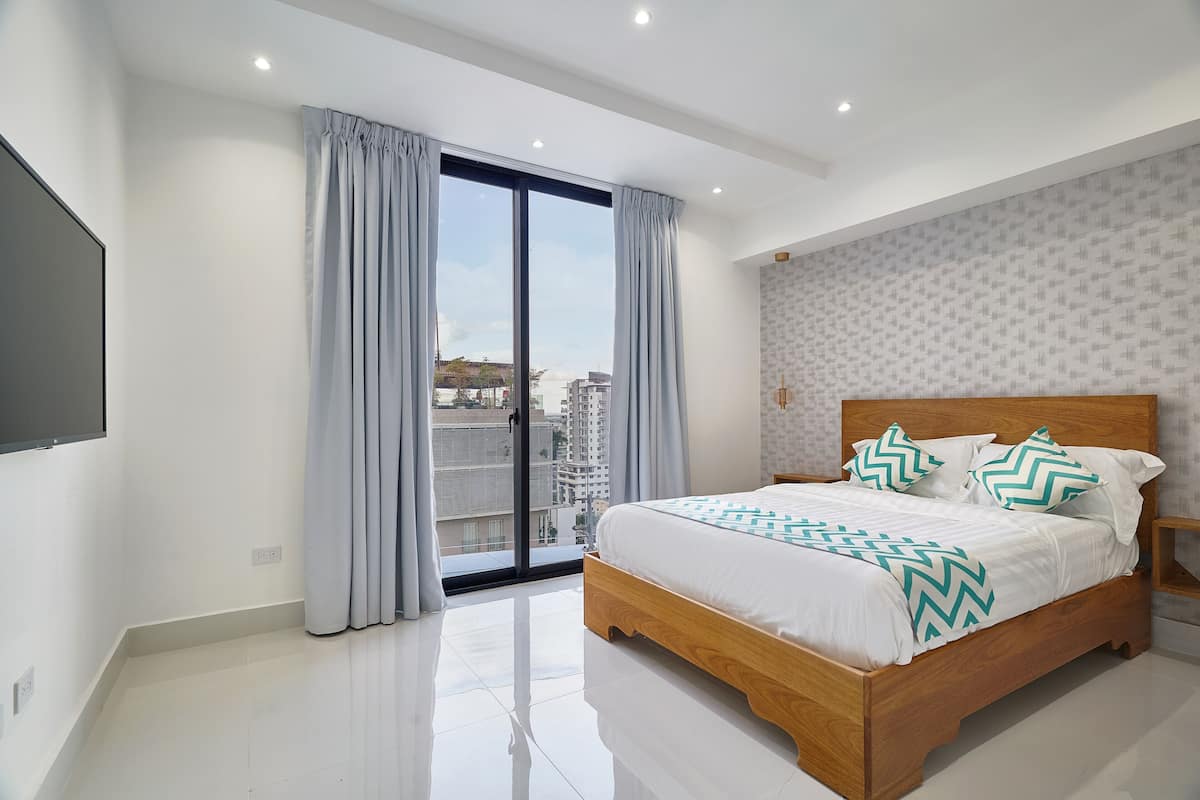 A bedroom with a large bed, a TV mounted on the wall, and a view of buildings through the window.