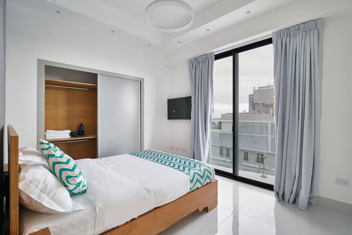 A modern bedroom with a bed, built-in wardrobe, a flat-screen TV, and a large window with curtains.