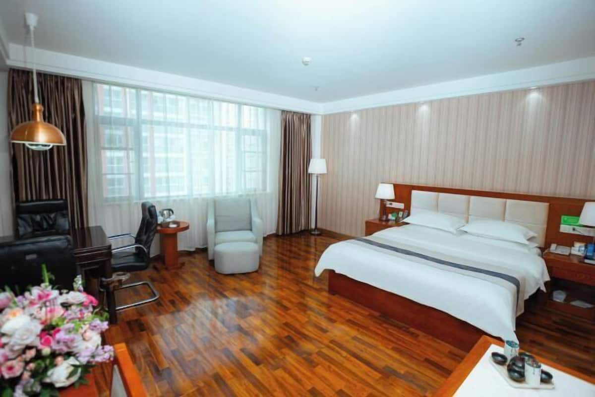 A hotel room with a wooden floor, a large bed, a desk with a lamp, a chair, and a window with curtains.