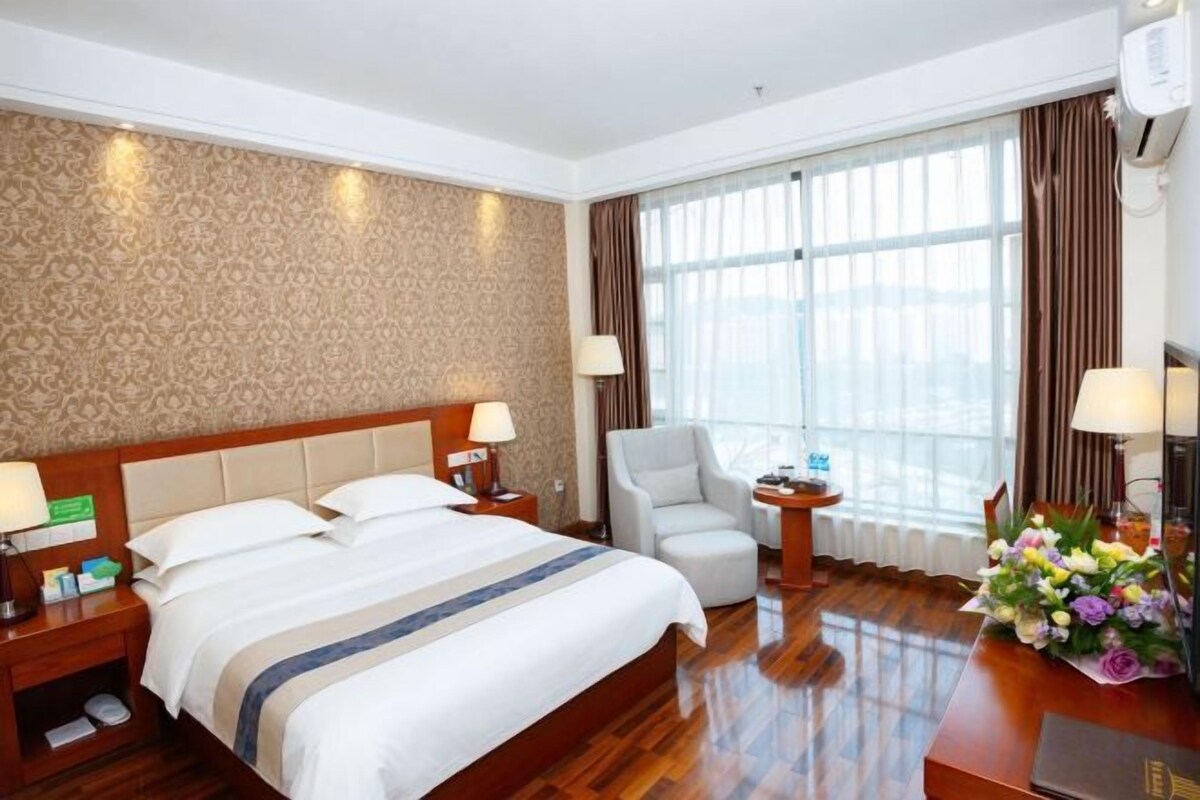 Habitación de hotel con una cama grande, un escritorio, una silla y vistas al exterior a través de amplias ventanas.