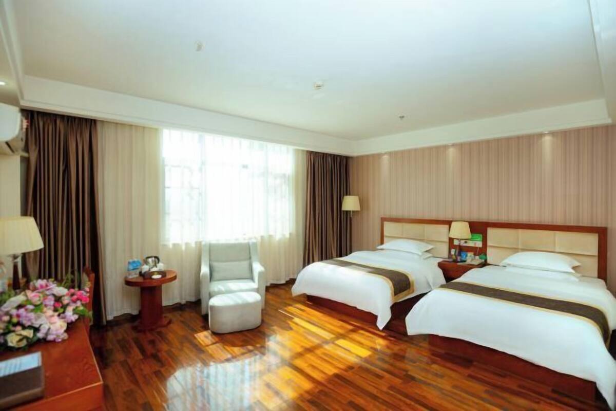 A hotel room with two beds, a sitting area with a chair, a small table, and a desk with a lamp.