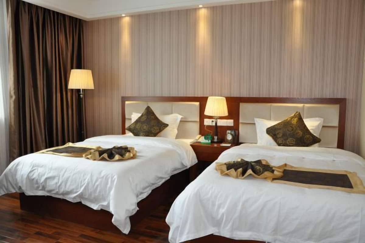 A hotel room with two beds, each with white linens and decorative pillows. There are two bedside lamps and a nightstand with a lamp and a clock. The room has wooden flooring and a curtain on one side.