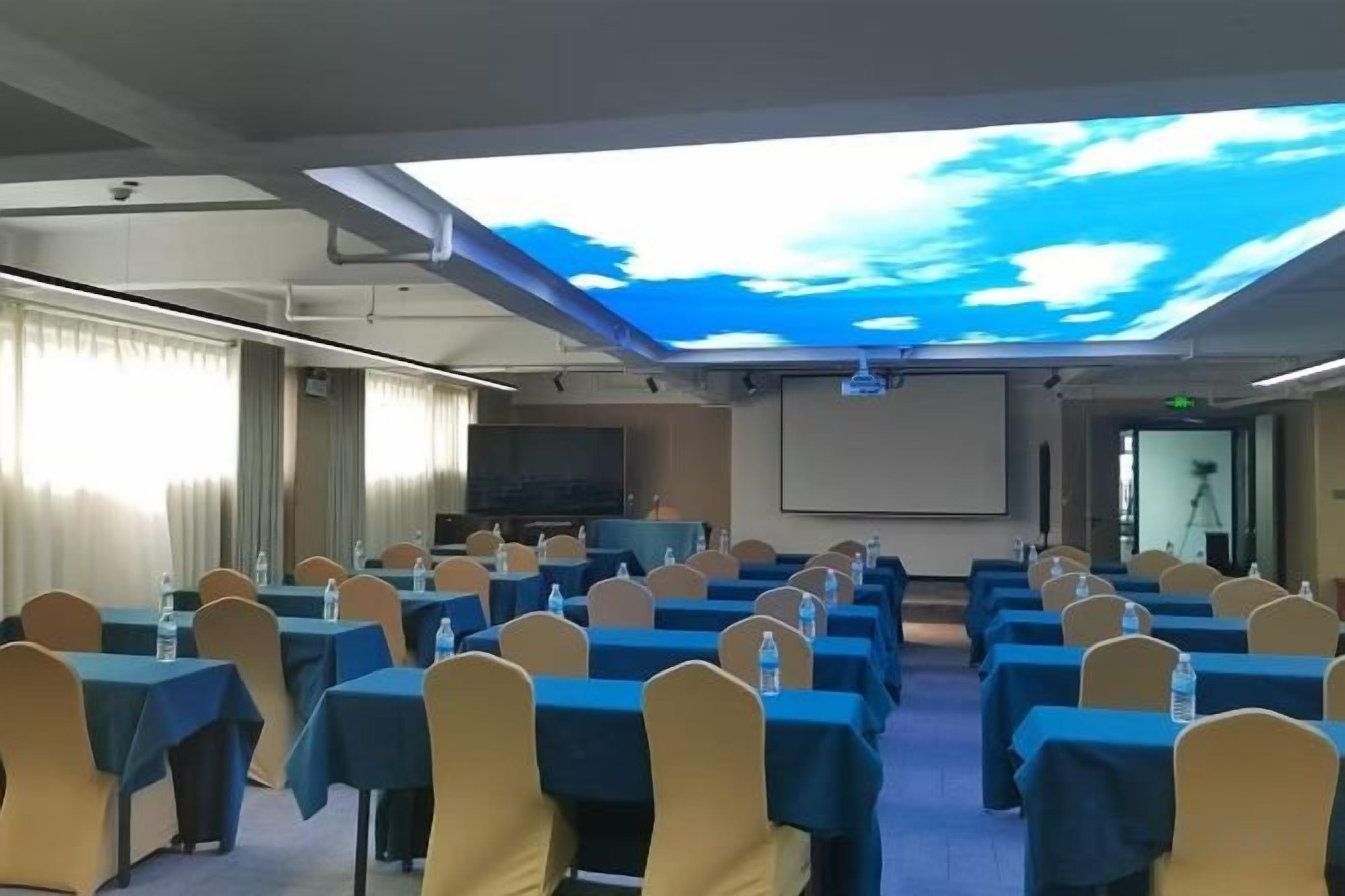Ein Konferenzraum mit Tischen, Stühlen, einer Projektionsleinwand und einem großen Bildschirm, der einen Himmel mit Wolken zeigt.