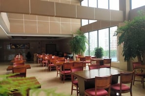 A spacious dining area with multiple tables and chairs, large windows, and potted plants.