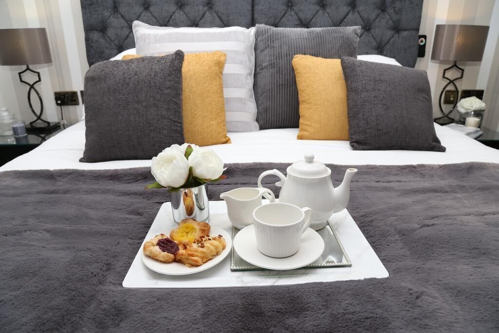 Una cama bien hecha con almohadas grises y mostaza, una bandeja con pasteles, un hervidor de agua y una taza sobre una colcha gris esponjosa.