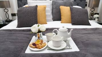 Una cama bien hecha con almohadas grises y mostaza, una bandeja con pasteles, un hervidor de agua y una taza sobre una colcha gris esponjosa.
