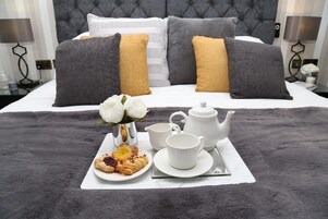 Una cama bien hecha con almohadas grises y mostaza, una bandeja con pasteles, un hervidor de agua y una taza sobre una colcha gris esponjosa.