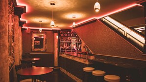 A dimly lit bar area with wooden tables and stools, a staircase, and a bar counter with shelves of bottles. - The Old Hall Hotel (Coventry)
