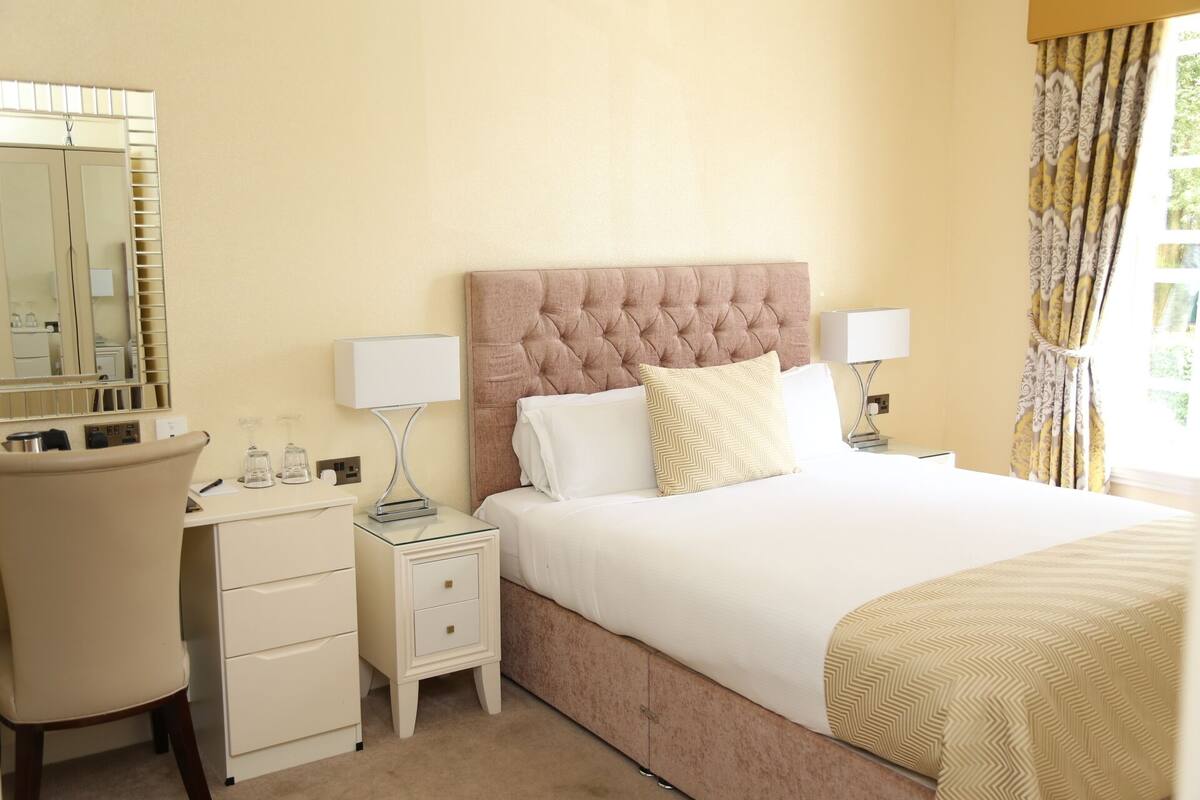A hotel room with a large bed, two bedside tables, a chair, a mirror, and a window with curtains.