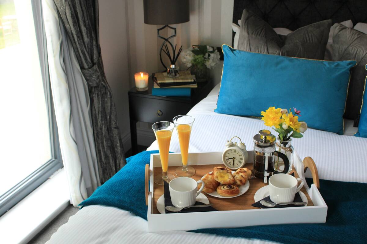 A neatly made breakfast tray with pastries, two glasses of orange juice, and a cup of coffee on a bed with a blue pillow and a nightstand with a book and a candle.