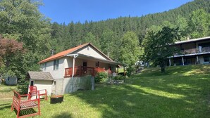 Ein zweistöckiges Haus mit Veranda und kleiner Terrasse, umgeben von einem grünen Rasen und einem bewaldeten Hang.