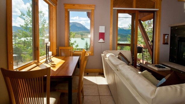 A cozy room with a wooden dining table, a sofa, and large windows offering a scenic view of mountains and a lake. - Mountain-View Condo in Dillon • Walk to Dining & Lake • Close to Keystone (Dillon)