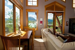 A cozy room with a wooden dining table, a sofa, and large windows offering a scenic view of mountains and a lake. - Mountain-View Condo in Dillon • Walk to Dining & Lake • Close to Keystone (Dillon)