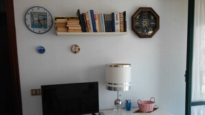 A room with a white desk, a black monitor, a lamp, a bookshelf with books, a clock, and a window with curtains.