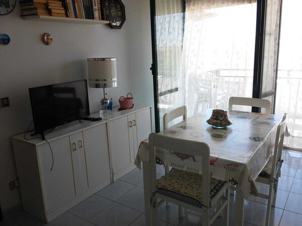 A dining area with a table covered in a tablecloth, chairs, a television, a lamp, and a basket on a cabinet.