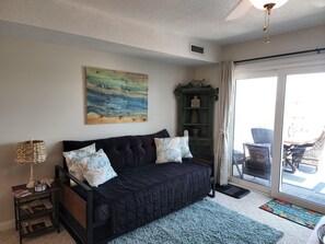 A cozy living room with a dark-colored sofa, a decorative painting, a wicker side table, and a view through a glass door.