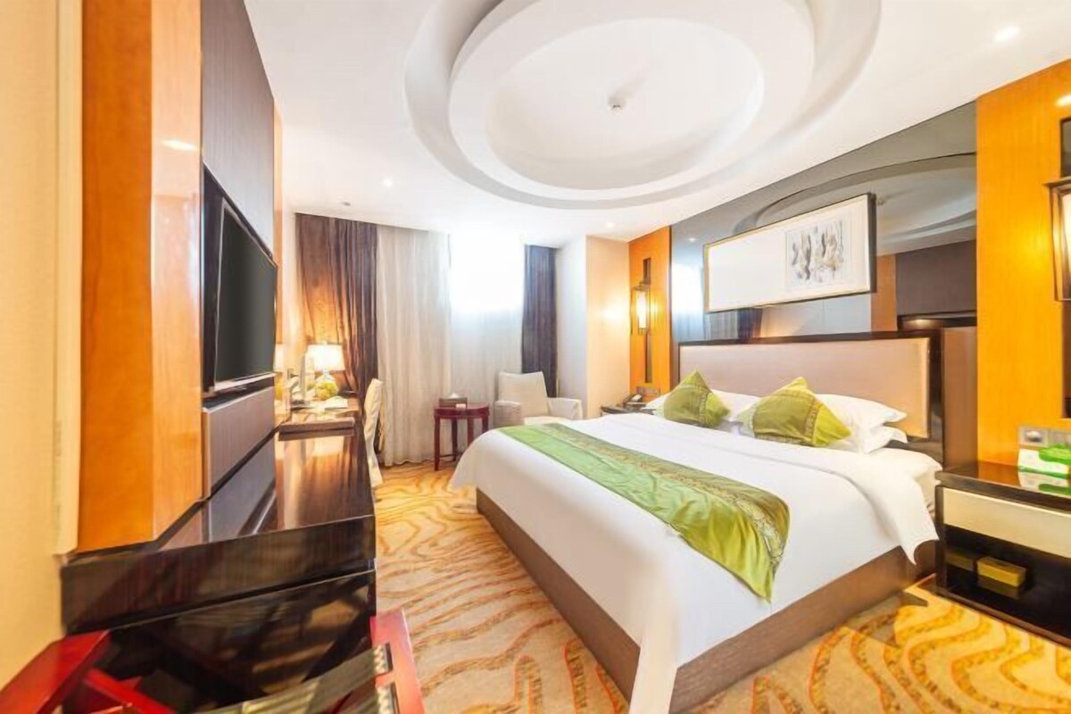 A hotel room with a large bed, a flat-screen TV, a desk with a lamp, and a small side table.