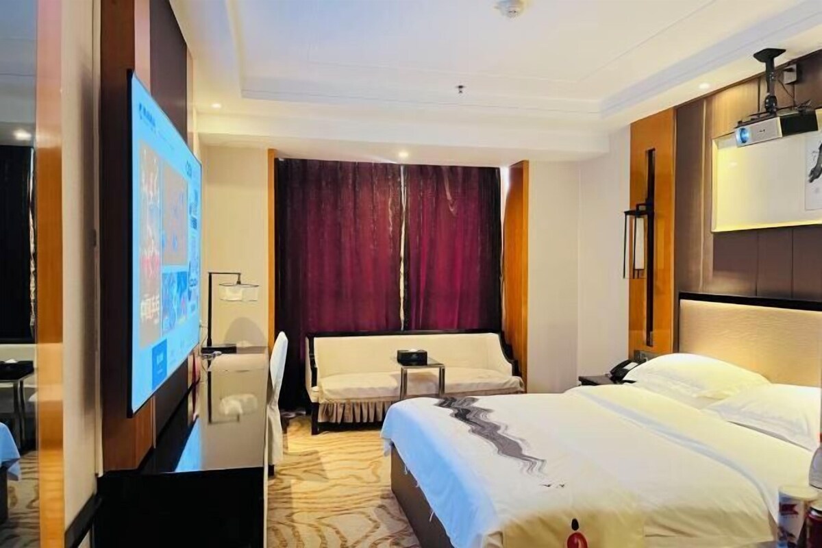 A hotel room with a bed, a desk with a computer, a TV, and a large window with curtains.