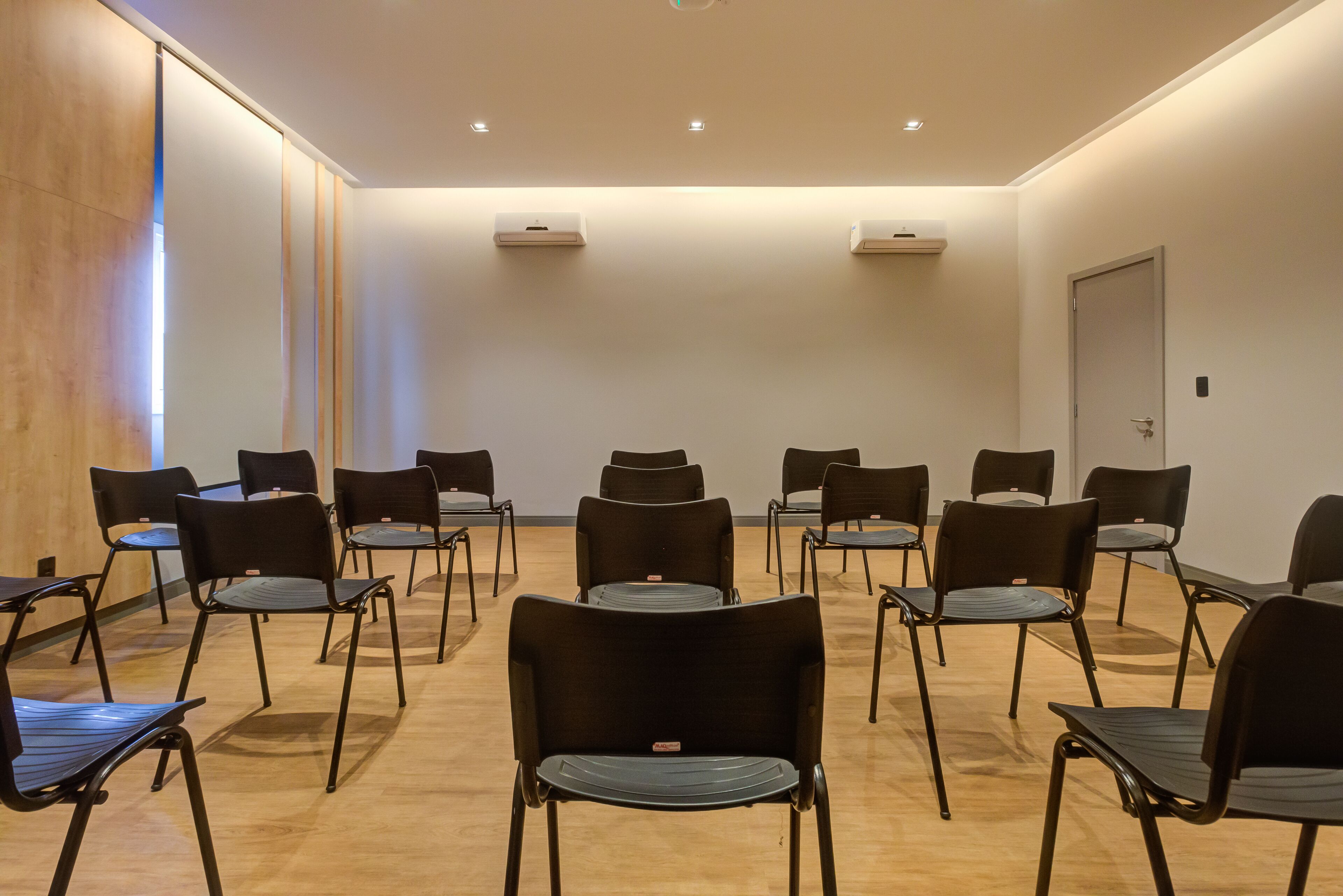 Sala de conferência com fileiras de cadeiras pretas, uma parede de destaque em madeira e iluminação embutida no teto.