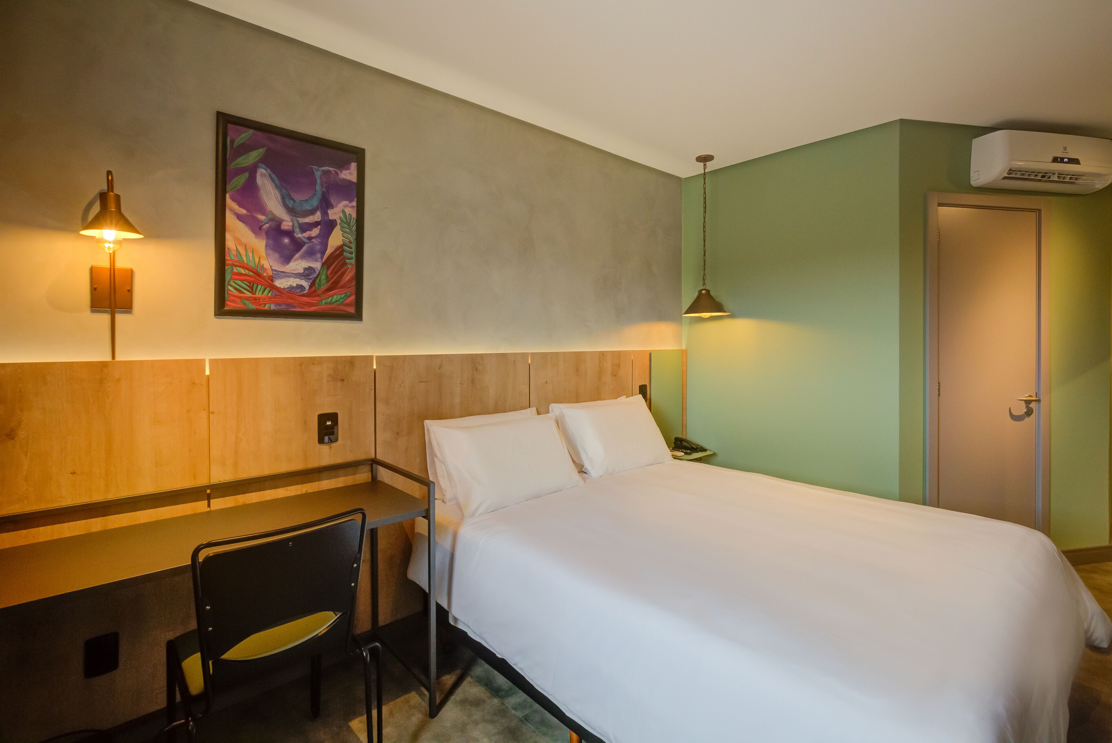 A hotel room with a bed, a desk, a chair, a painting on the wall, and an air conditioning unit.