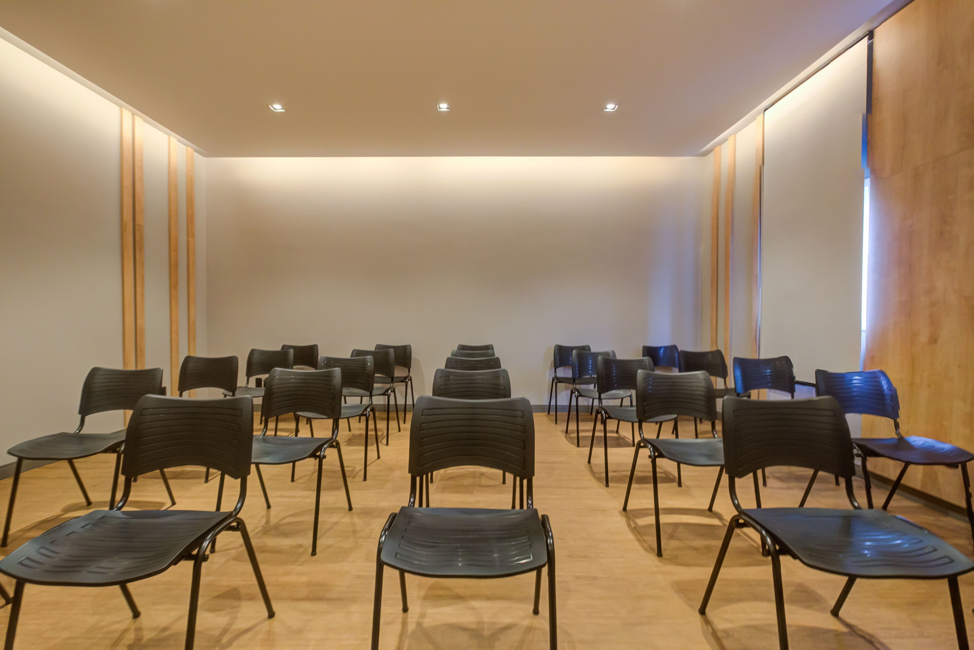 Un salón de conferencias con filas de sillas y mesas, paneles de madera en las paredes y luces empotradas en el techo.