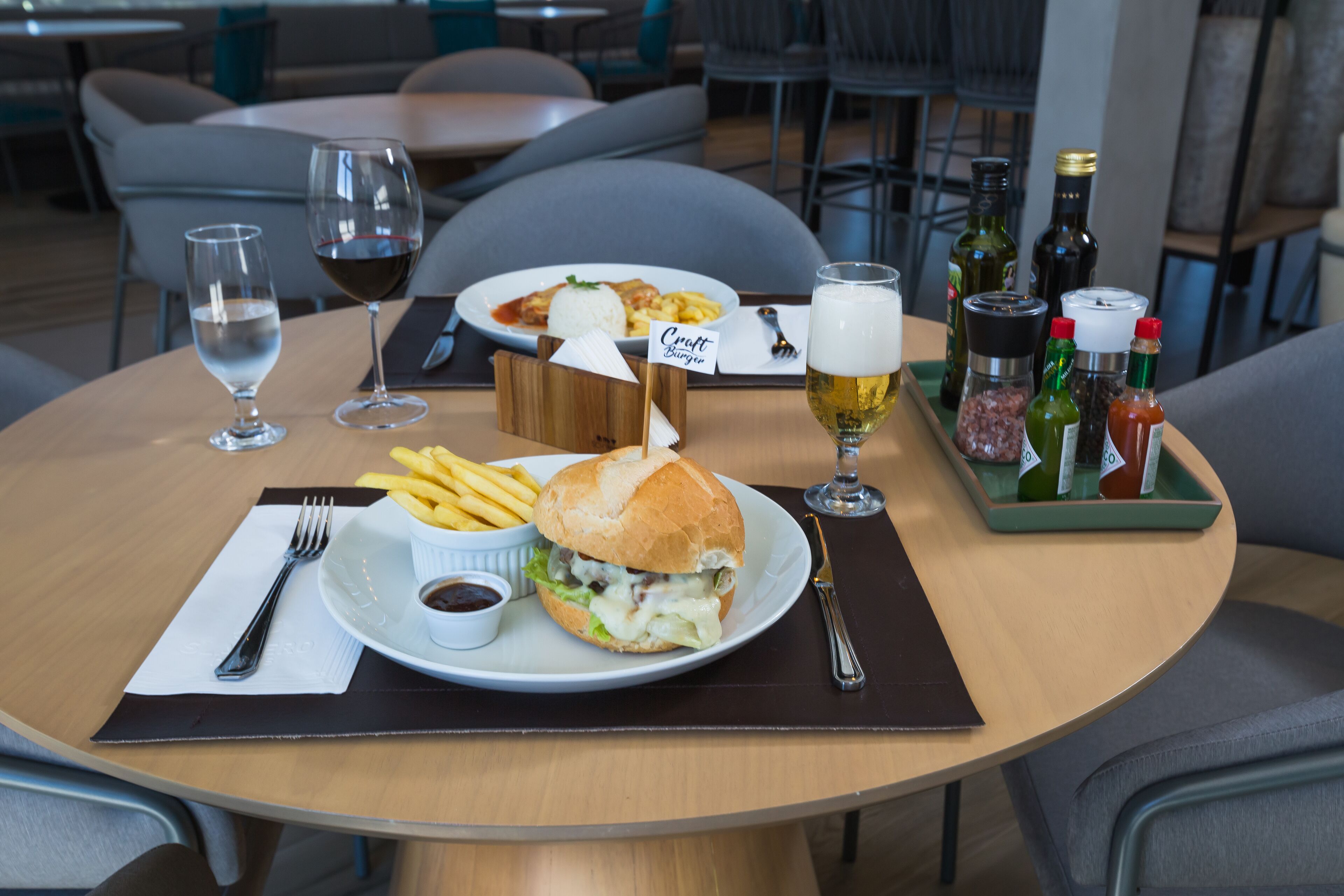 Una mesa de comedor con una comida que incluye una hamburguesa, papas fritas y un pequeño bol de salsa. También hay dos copas de vino y una bandeja con condimentos.